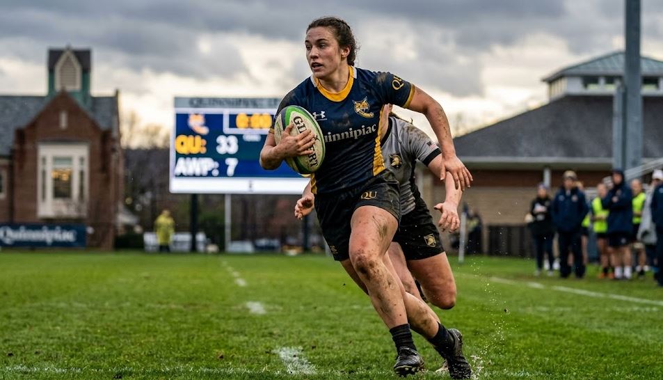 Quinnipiac Women's Rugby player running with the ball during a Rugby 7s match, with a scoreboard showing a 33-7 victory over Army West Point