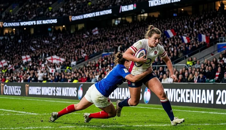 A realistic action photograph capturing an England Red Roses player driving forward with the rugby ball during an evening match at Twickenham. She is being tackled low by a France player in a packed stadium with "GUINNESS WOMEN'S SIX NATIONS 2026" displayed on the pitchside LED boards. Sweat and determination are visible.