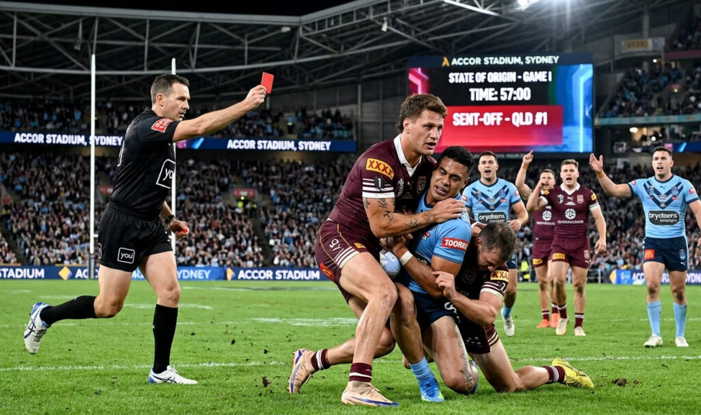 A high-resolution 4k photograph of Queensland Maroons star Kalyn Ponga (using face reference from original) performing a high-contact tackle on a New South Wales Blues player in State of Origin Game I, resulting in a direct red card. Ponga (maroon jersey #1) has his arm around the head/neck area of the player in blue. The referee in the foreground is holding up a clear red card. The scoreboard on the jumbotron in the distance confirms the time as 57:00 and the 'sent-off' status, with other players from both teams reacting to the decision. Scene at Accor Stadium, Sydney.