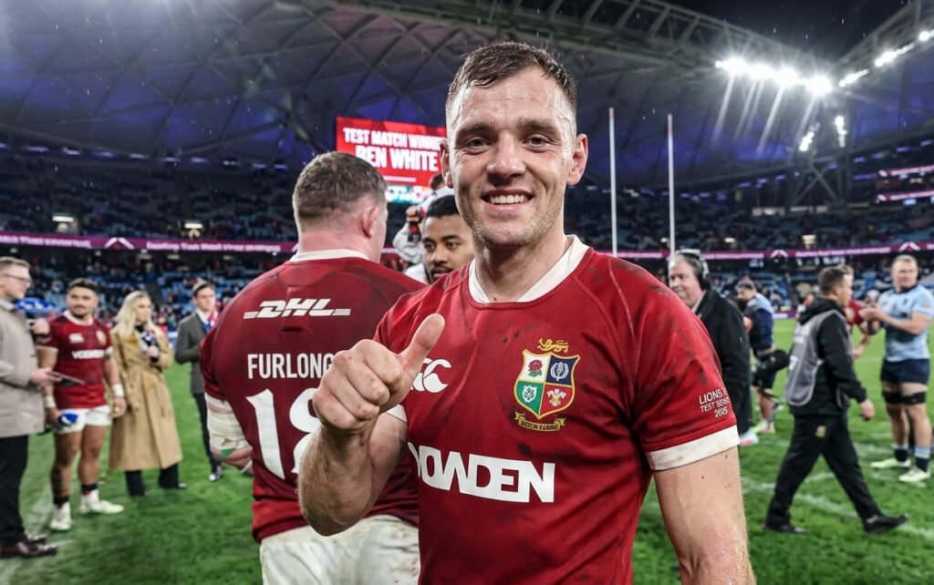 Ben White smiling and giving a thumbs-up on the pitch after a match, wearing the red British & Irish Lions jersey with a packed stadium and bright floodlights in the background.