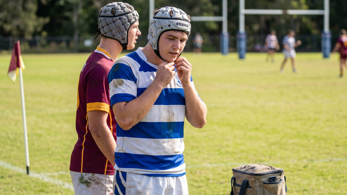 A candid, realistic photograph of two Australian rugby players on a sideline, wearing different styles of rugby headgear (one open-ear, one full-coverage), looking onto a grassroots rugby pitch during bright daylight.