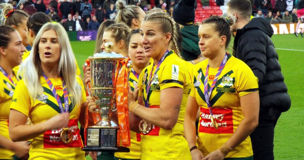 Australian women's rugby league players in gold jerseys holding the World Cup trophy