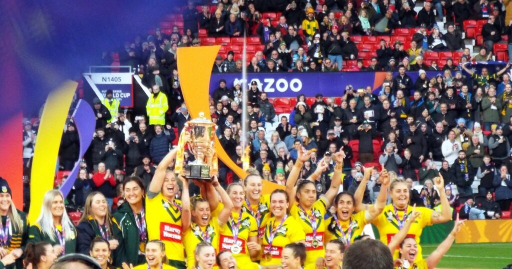 Australian women's rugby league players in yellow lifting the World Cup trophy in front of a crowd