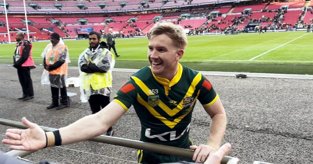 Tom Dearden in a green and gold Kangaroos jersey smiling and reaching towards fans at Wembley